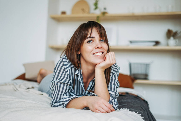 Молода щаслива брюнетка в повсякденному одязі відпочиває на ліжку в яскравому інтер'єрі
  - Фото, зображення