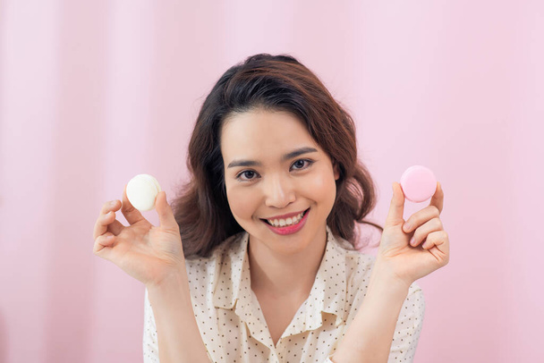 Joven mujer asiática alegre sosteniendo dos macarons delante de ojos sobre fondo rosado
. - Foto, imagen