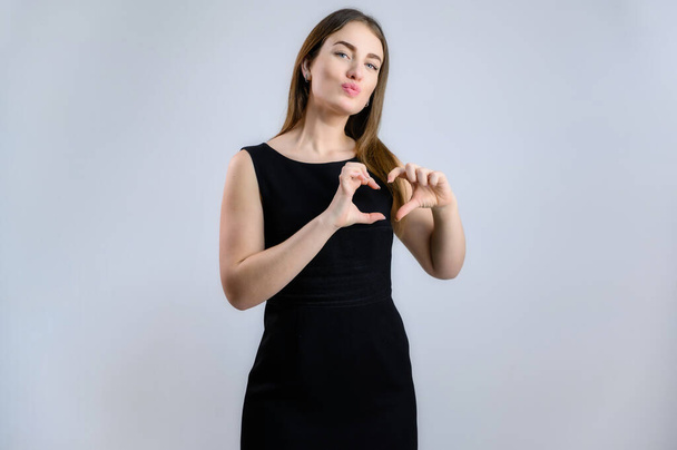 Portrait on a white background of a sympathetic Caucasian woman in a black dress shows fingers a love heart with a smile - Fotó, kép