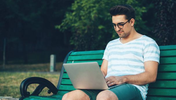 Белый мужчина в очках сидит на скамейке в парке и пользуется ноутбуком.
 - Фото, изображение