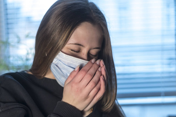 Πρόληψη της εξάπλωσης της νόσου του Coronavirus 2019 (Covid-19). Νεαρή γυναίκα φτερνίζεται ή βήχει σε προστατευτική μάσκα προσώπου  - Φωτογραφία, εικόνα