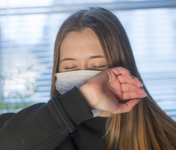 Πρόληψη της εξάπλωσης της νόσου του Coronavirus 2019 (Covid-19). Νεαρή γυναίκα φτερνίζεται ή βήχει σε προστατευτική μάσκα προσώπου - Φωτογραφία, εικόνα
