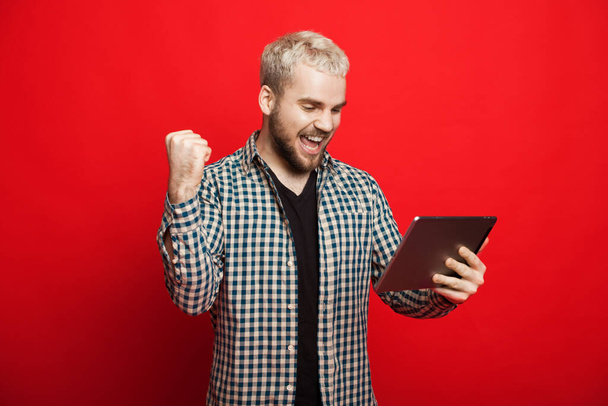 Caucásico hombre con el pelo rubio y la barba es un gesto de ganar mientras sostiene una tableta y posando sobre un fondo rojo
 - Foto, imagen