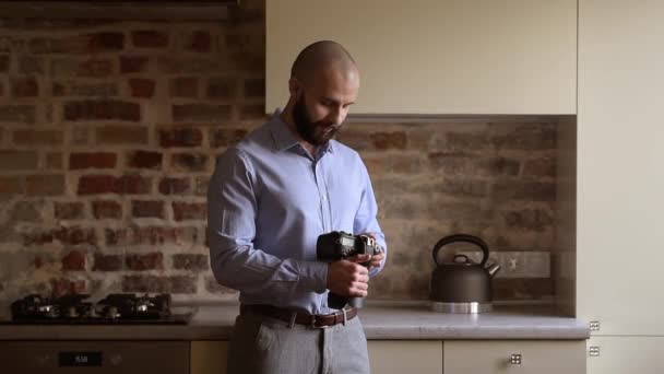 A bald photographer searches for something in his camera in the dining room. A man with a beard wears two silver earrings, a blue shirt, grey trousers, and a brown leather belt. - Materiał filmowy, wideo