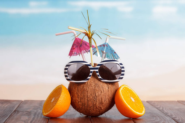 Coconut with straw, umbrellas, sunglasses and a part of orange on a brown table against the sea. - 写真・画像