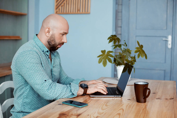 Un hombre atractivo adulto trabaja en la computadora durante el café de la mañana. Hombre de negocios escribiendo en un portátil durante el desayuno. Negocios, trabajo, trabajo en casa
 - Foto, imagen