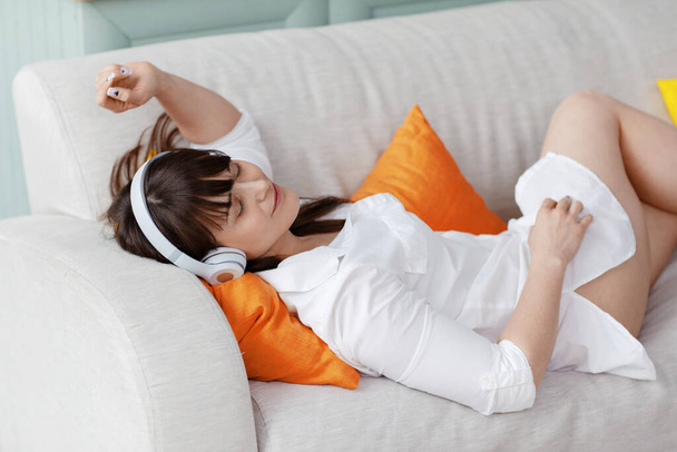 Ένα νεαρό όμορφο κορίτσι με λευκό πουκάμισο απολαμβάνει τη μουσική ενώ ξαπλώνει σε έναν καναπέ ανάμεσα σε φωτεινά μαξιλάρια. Ξεκούραση, χαλάρωση, τρόπος ζωής - Φωτογραφία, εικόνα
