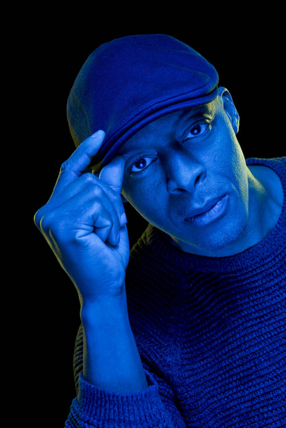 Black man with blue and yellow light, with hand on the flat cap, isolated on black background, looking at the camera. Greeting concept. Vertical - 写真・画像