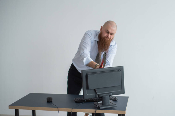 Angry bald man with a red beard in the office in a business suit smashes an ax with a computer. The manager with a nervous breakdown, breaks the monitor. - Valokuva, kuva