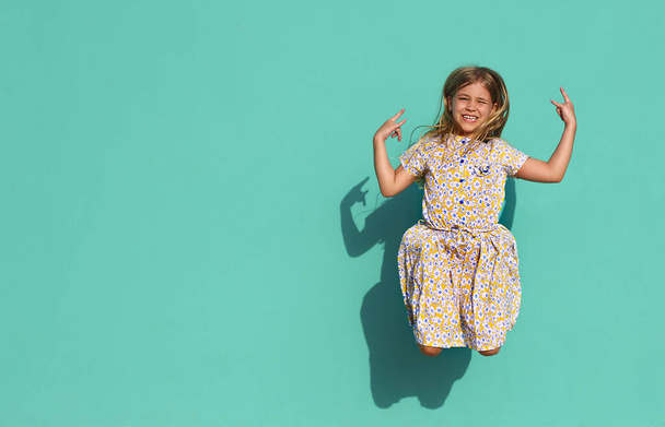 Весела маленька дівчинка в барвистій сукні показує козячий жест і стрибає проти зеленої стіни на сонячному світлі
 - Фото, зображення