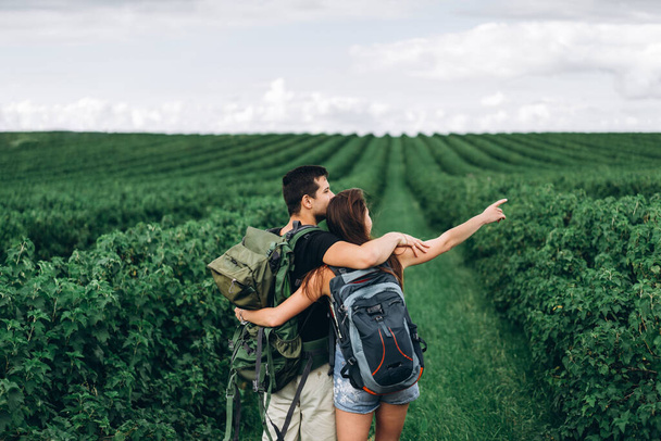 Back view of young couple with backpacks on currant plantations. Woman with long hair points her hand to the horizon. - Фото, изображение