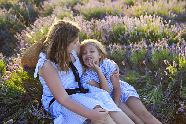 Van boven jong vrouwelijk en klein meisje in blauw geruite jurken omhelzen terwijl ze samen zitten in lavendelveld in de zonnige zomerdag - Foto, afbeelding