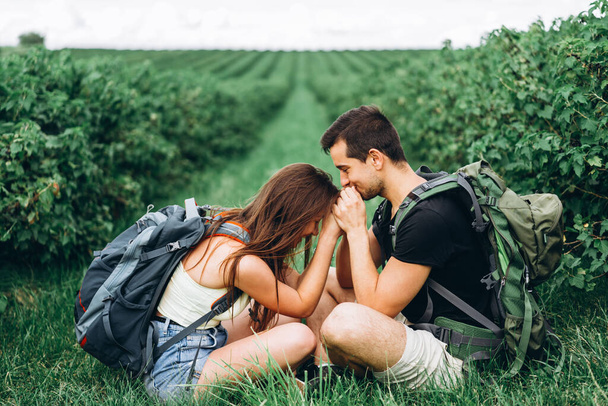 Man and woman with backpacks on their backs sitting hugging on the green grass between the currant bushes. Walking on the currant plantations in the countryside. - Foto, immagini