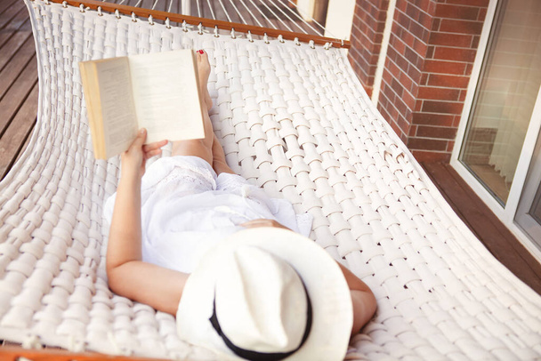 Desde arriba de mujer joven irreconocible en vestido blanco y sombrero acostado en hamaca y libro de lectura mientras se relaja en la terraza de la casa
 - Foto, Imagen