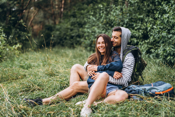 Woman with long hair and man sitting on the grass in the forest with backpacks. Have fun in nature, hugging and kissing. - Foto, Imagem
