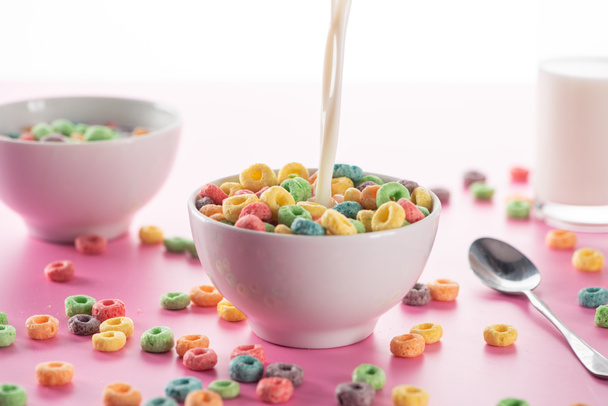 selective focus of bright multicolored breakfast cereal in bowl with pouring milk near spoon on pink background - Photo, image