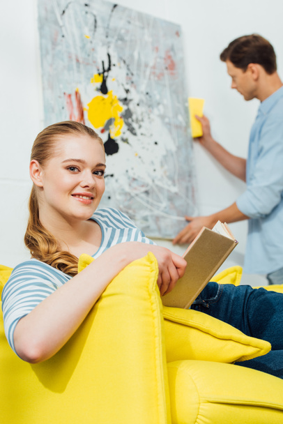 Selective focus of girl with book smiling at camera near boyfriend cleaning painting in living room - Foto, afbeelding