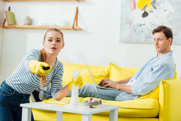 Focus selettivo della donna con prodotti per la pulizia utilizzando telecomando vicino al fidanzato con laptop sul divano a casa
 - Foto, immagini