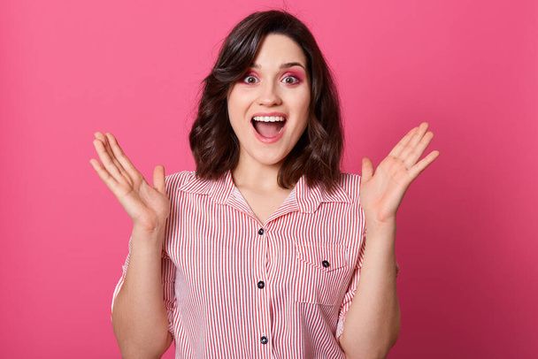 Photo of emotional magnetic charming young brunette opening mouth and eyes widely, raising both hands, looking directly at camera, standing isolated over pink background, being in high spirits. - Foto, immagini