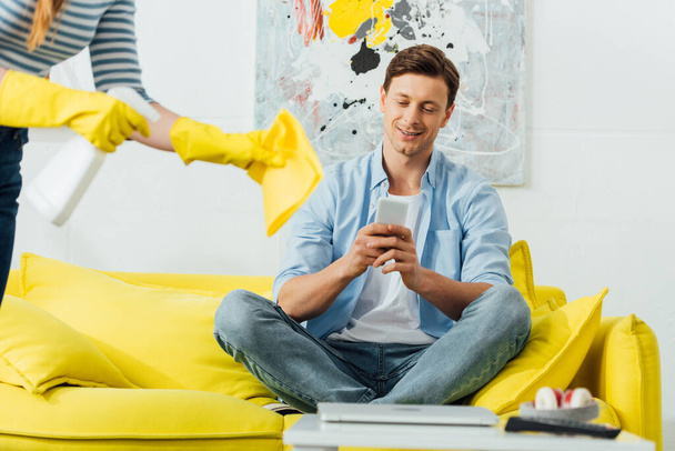 Selektiver Fokus eines lächelnden Mannes mit Smartphone auf Couch neben Frau mit Waschmittel und Lappen im Wohnzimmer - Foto, Bild