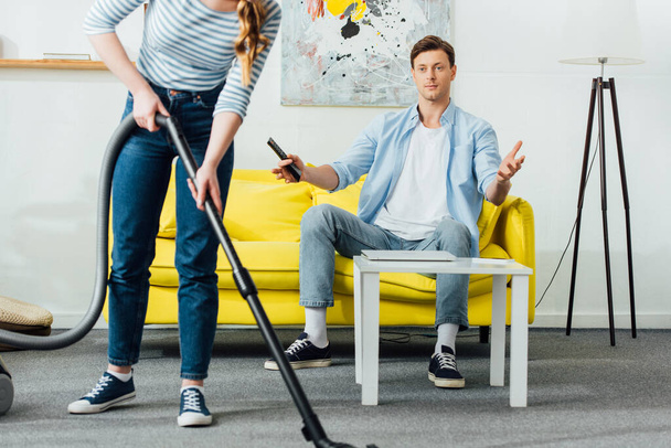Женщина чистит ковер рядом удивленный парень с пультом дистанционного управления на диване дома
 - Фото, изображение