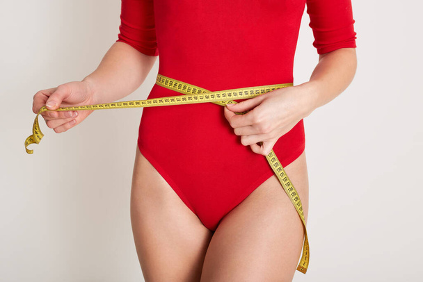 Indoor picture of unknown female body measuring her waist with yellow measuring tape, caring about her body, keeping fit, losing weight, being on diet, wearing red combidress. Women and diet concept. - Foto, afbeelding