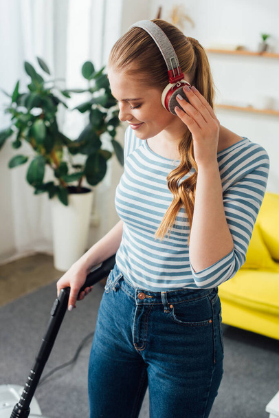 Mujer sonriente escuchando música en auriculares mientras limpia la alfombra con aspiradora en casa
  - Foto, imagen