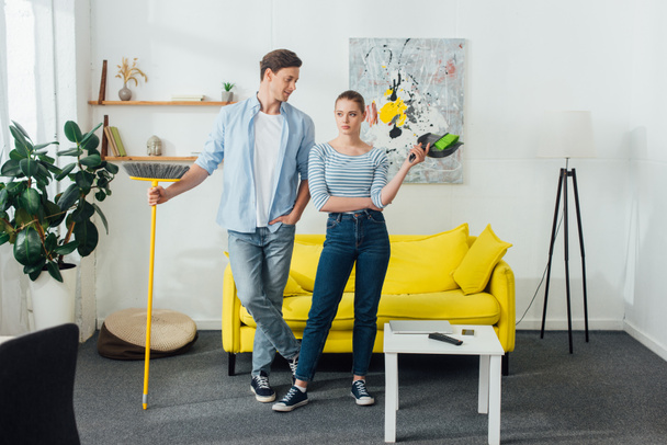 Man looking at girlfriend with scoop and broom in living room - Foto, Imagem