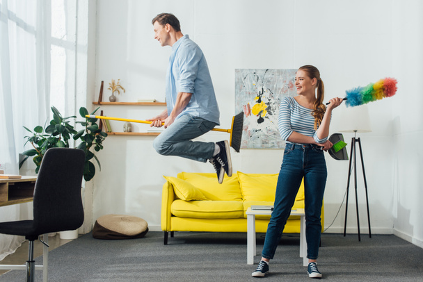 Smiling woman with dust brush and scoop looking at boyfriend levitating on broom in living room - Foto, imagen
