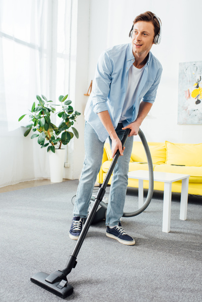 Smiling man cleaning carpet with vacuum cleaner and listening music in headphones at home  - Valokuva, kuva