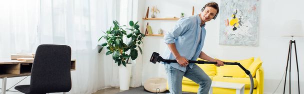 Side view of cheerful man in headphones holding brush of vacuum cleaner in ling room, panoramic shot - Fotoğraf, Görsel