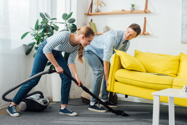 Side view of smiling woman cleaning carpet with vacuum cleaner near man lifting up couch at home - Foto, Bild