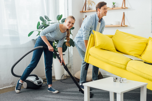 Smiling man lifting up couch near girlfriend with vacuum cleaner in living room - Photo, Image
