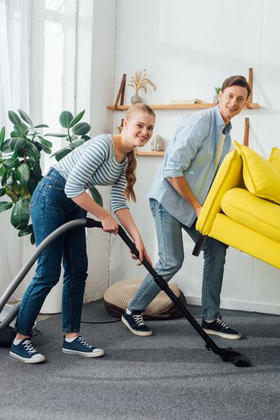 Smiling couple looking at camera while cleaning carpet with vacuum cleaner under couch in living room - Foto, Imagen