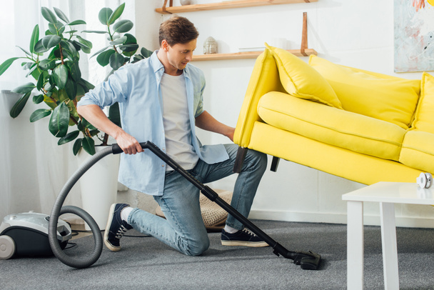 Side view of man lifting up couch while cleaning carpet with vacuum cleaner  - Photo, Image