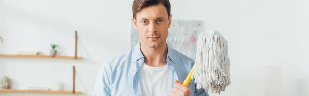 Handsome man looking at camera and holding mop in living room, panoramic shot  - Foto, Imagen