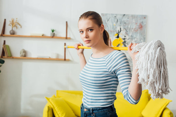 Beautiful woman looking at camera while holding mop at home - Fotoğraf, Görsel