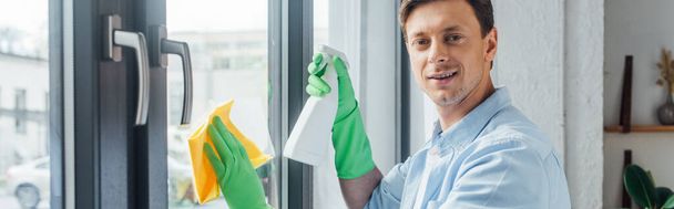 Seitenansicht eines Mannes mit einer Flasche Waschmittel und einem Lappen, der in die Kamera lächelt, während er zu Hause Fenster putzt, Panoramaaufnahme - Foto, Bild