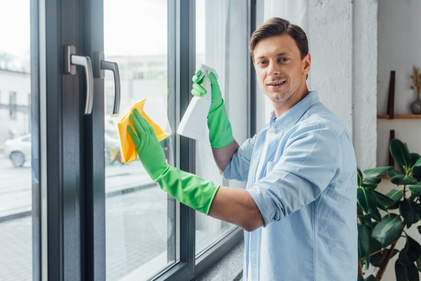 Вид сбоку человека с бутылкой моющего средства и тряпкой, улыбающегося на камеру во время мытья окон дома
 - Фото, изображение