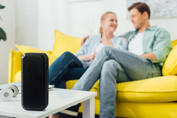 Selective focus of wireless speaker and headphones on coffee table near young couple on couch at home - Foto, Bild
