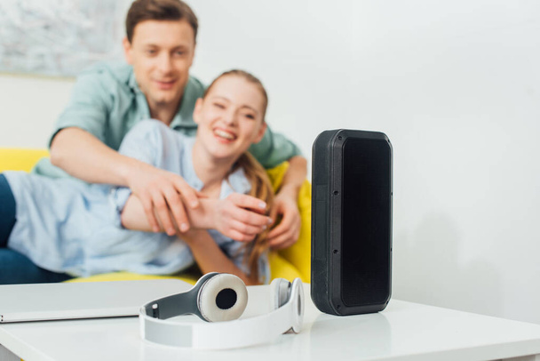 Selective focus of wireless speaker, headphones and laptop on coffee table near cheerful couple on couch at home - Photo, Image