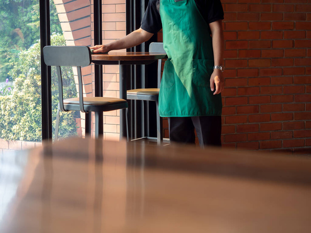 Camarero con delantal verde limpiando la mesa redonda de madera cerca de la ventana de vidrio y la pared de ladrillo en la cafetería de estilo loft moderno con espacio para copiar
. - Foto, imagen