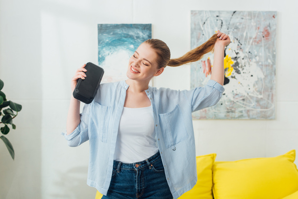 Cheerful woman touching hair while holding portable speaker in living room - Photo, Image