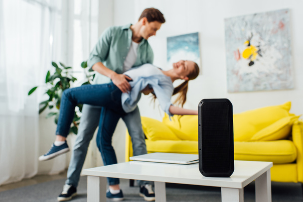 Selective focus of wireless speaker and laptop on coffee table and smiling young couple dancing at home - Φωτογραφία, εικόνα