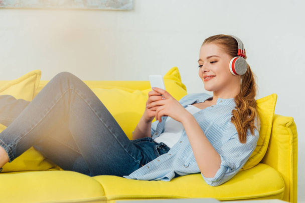 Selective focus of smiling woman in headphones using smartphone on couch in living room - Fotó, kép