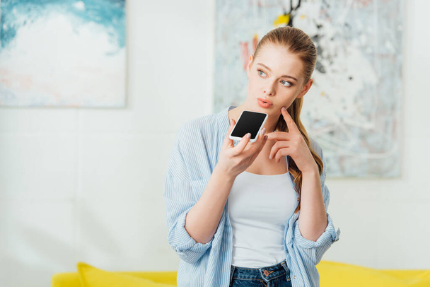 Menina pensativa com o dedo perto da bochecha falando no smartphone em casa
  - Foto, Imagem