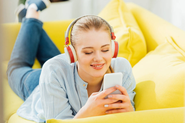 Selective focus of smiling girl in headphones using smartphone on couch  - Foto, Imagen