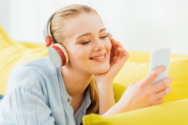 Selective focus of smiling woman in headphones using smartphone on sofa  - Photo, Image