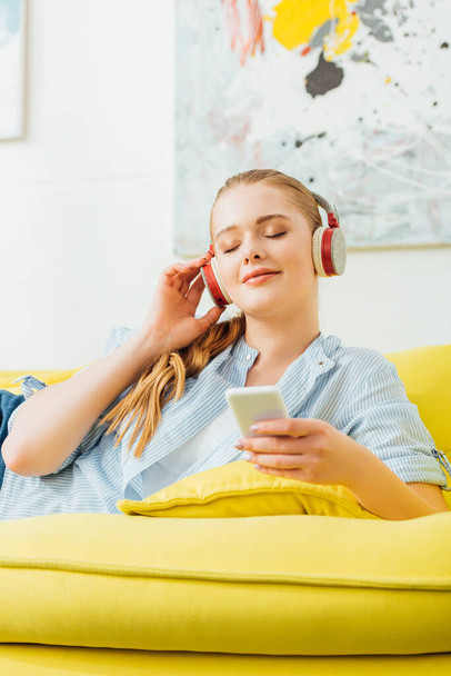 Attractive girl with closed eyes listening music in headphones and holding smartphone on couch  - Zdjęcie, obraz