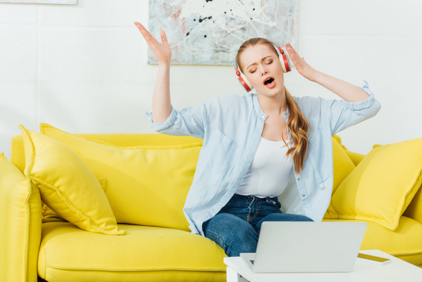 Attraktive Frau singt beim Musikhören über Kopfhörer auf Sofa neben Laptop und Smartphone auf Couchtisch  - Foto, Bild